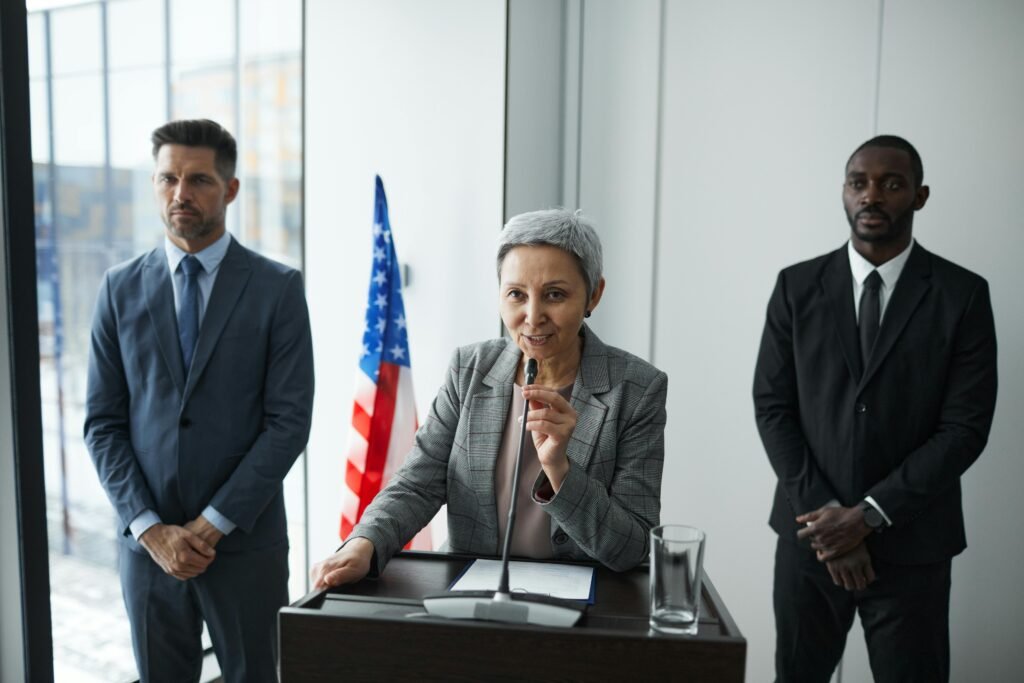 A female politician delivers a speech with bodyguards and an American flag in the background.